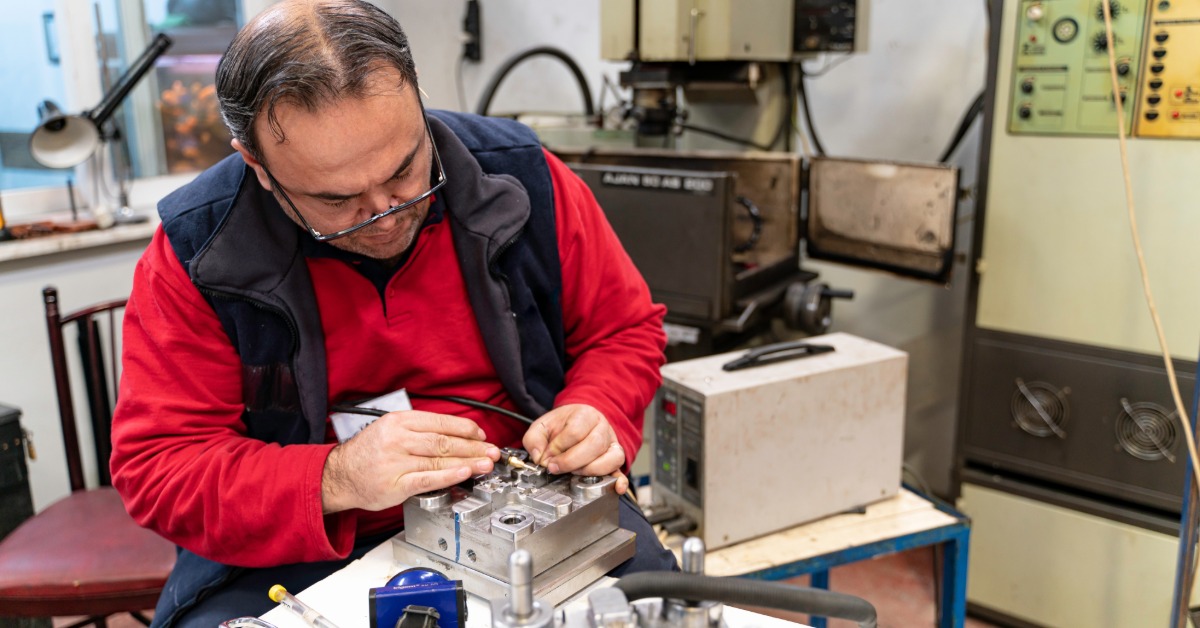 A man wearing a red shirt, black vest, and glasses is using a tool to work on a metal mold. He's sitting at a table.
