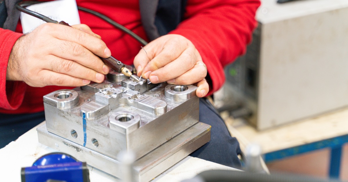 A close-up view shows a man wearing a red shirt and black vest using a tool to work on a metal mold.